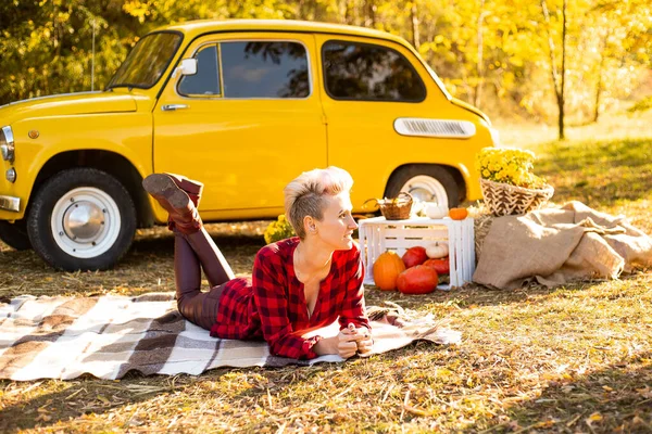 Sonbahar parkında rahatlama, güz parkında battaniyeye uzanmış sıradan giyinmiş mutlu hippi kadın sarı arabanın, çiçeklerin ve balkabaklarının yanında.