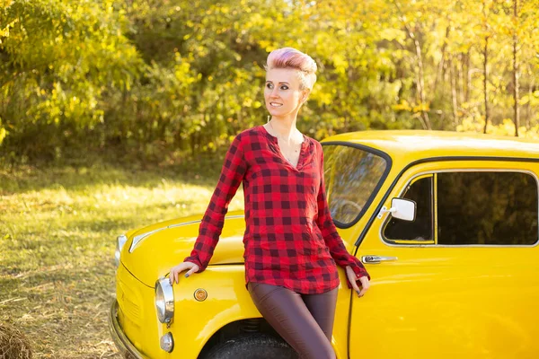 Sonbahar parkında sarı arabanın yanında kısa saçlı, altın ağaçlı bir kadın.
