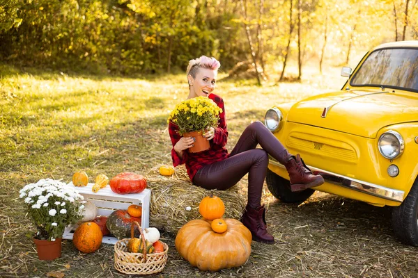 Sonbahar parkında sarı arabanın yanında çiçekli ve balkabaklı mutlu bir kadın.