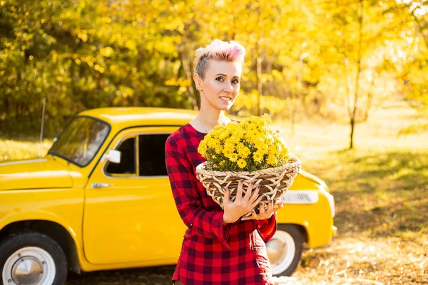 Sonbahar parkında sarı arabanın yanında çiçekli ve balkabaklı güzel bir kadın sepeti.
