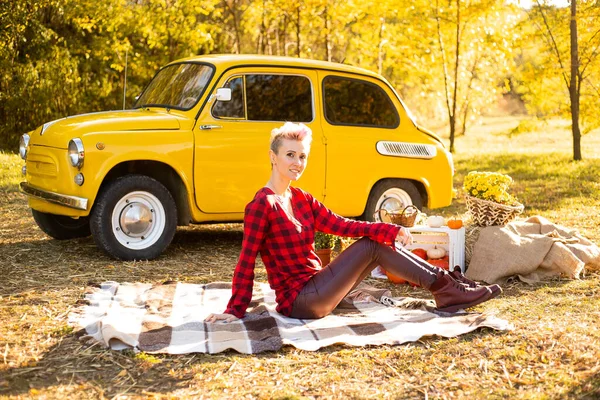Sonbahar parkında dinlenmek, gündelik giysiler içindeki hippi kadın sonbahar parkında arka planda, sarı arabanın, çiçeklerin ve balkabaklarının yanında battaniyeye uzanmak.