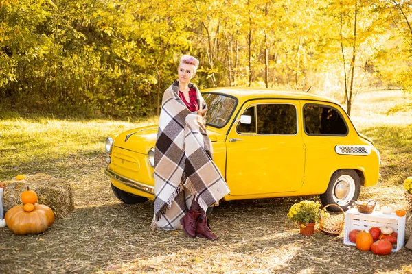 Güz çiçekleri arka planında altın ağaçlarıyla sarı retro arabanın yanındaki battaniyeye sarılı kısa saçlı şık hippi kadın.