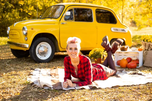 Sonbahar parkında, arka planda, sarı arabanın, çiçeklerin ve balkabaklarının yanında battaniyeye uzanmış bir kadın.