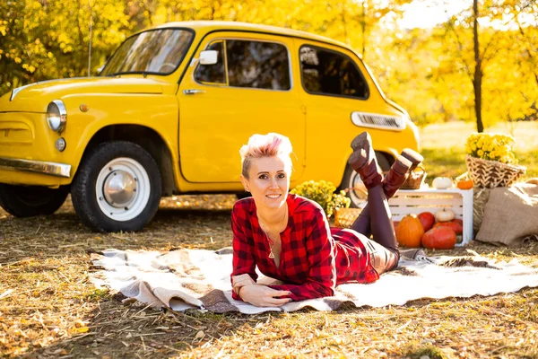 Sarı arabanın, çiçeklerin ve balkabaklarının yakınındaki sonbahar parkında battaniyeye uzanmış kısa saçlı mutlu hippi kadın.