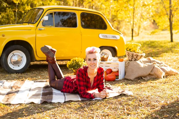 Sonbahar parkında rahatlama, sonbahar parkında, sarı araba, çiçekler ve balkabaklarının yanında, gündelik giysiler içinde battaniyeye uzanan hippi kadın.