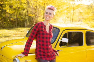 Sonbahar parkında sarı arabanın yanında kısa saçlı, altın ağaçlı hippi bir kadın.