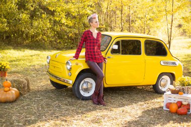 Sonbahar Park arka planında, sarı retro arabanın yanında kısa saçlı, altın ağaçlı hippi bir kadın.