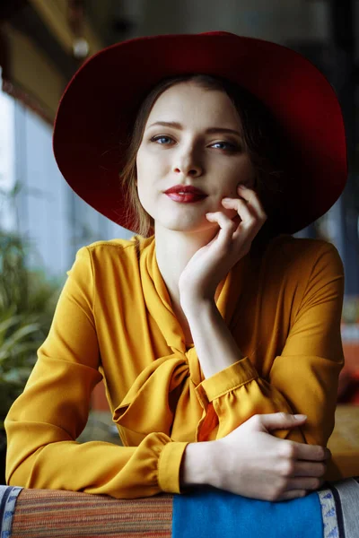 Retrato de una chica en un café con una taza de café y un sombrero.retrato de una chica joven sensual con sombrero flojo y blusa con lazo. Hermosa mujer morena en el café sosteniendo la taza de café —  Fotos de Stock