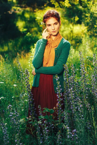 밭의 꽃들 사이에 갈색 머리카락을 가진 아름다운 소녀의 초상화. 여름 방학에 꽃이 핀다. 시골 스톡 사진