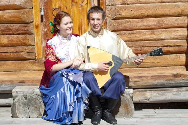 Menina bonita nova e o cara com a balalaika em traje nacional russo sentado no alpendre de uma casa de madeira — Fotografia de Stock