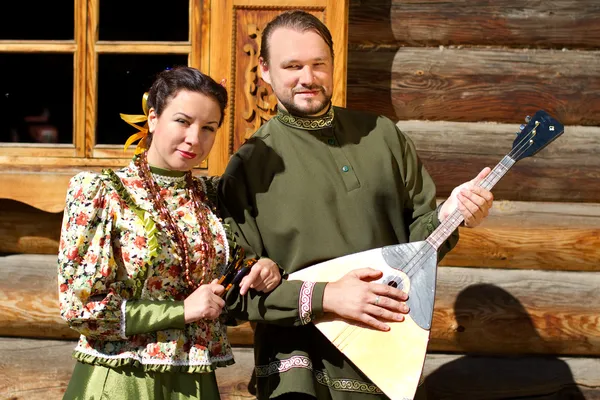 Une fille et un jeune homme avec balalaika cosaque en costumes nationaux contre une maison en bois — Photo