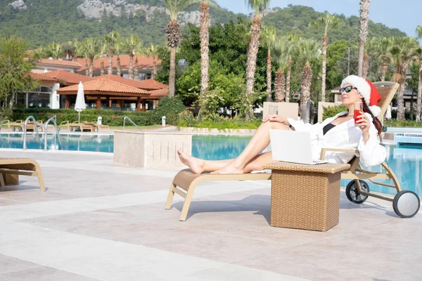 Yeni yıl şapkası ve beyaz önlüklü bir kız havuz kenarında dizüstü bilgisayar ve kokteyl ile oturuyor. Sıcak ülkelerde tatilde olan yeni yılı kutlama kavramı.