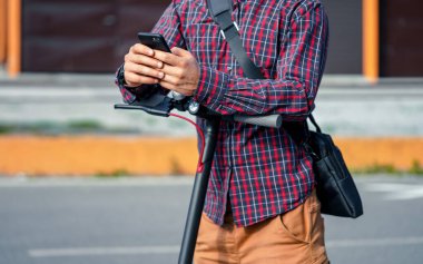 Genç adam, elektrikli scooter gidonuna yaslanmış, elinde cep telefonu tutan, yakın çekim detaylarıyla...