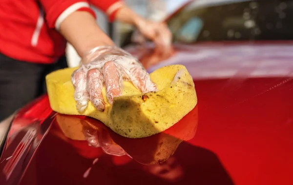 Young woman cleaning front hood of her car, closeup detail on yellow sponge held by hand in plastic glove — Stock Photo, Image