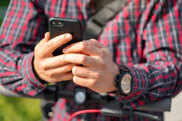 Giovane uomo che indossa una camicia appoggiata al manubrio dello scooter elettrico, tenendo in mano lo smartphone mobile, dettaglio primo piano — Foto Stock
