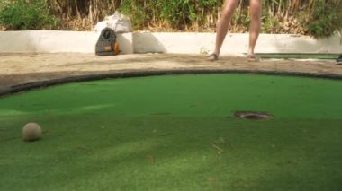 İnsan minigolf oynar, detaylar sadece ayaklarında kahverengi sandaletlerle yeşil yüzey üzerinde hareket eder ve beyaz top deliğe doğru hareket eder, onu eliyle çıkarır.