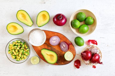 Avokado yarımları, parçalar, limonlar ve soğanlar, tuzlu fincan - beyaz çalışma tahtasında temel guacamole malzemeleri, düz yatar fotoğraf