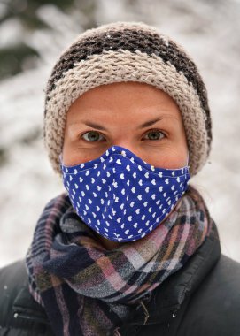 Kışlık ceketli, kalın şapkalı, pamuk virüsü maskeli, küçük kalpli, karla kaplı arka planda kalan ağaçlar, yakın plan detayları.