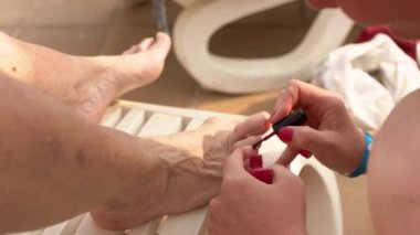Genç bir kadın yaşlı kadın ayaklarına kırmızı oje sürüyor. Ağır nasırlı. Ayrıca hallux valgus olarak da bilinir.