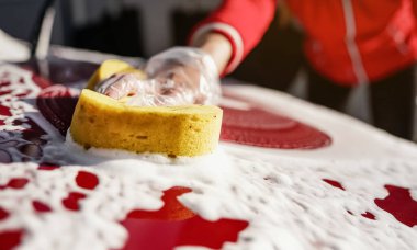 Arabasının ön kaputunu temizleyen genç bir kadın. Plastik eldivenle tutulan sarı süngerin yakın çekim ayrıntıları.