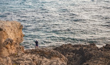 Tek bir balıkçı denizin kayalık sahilinde dikiliyor, arkadan bakıyor.