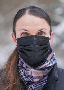 Siyah tek kullanımlık ceketli genç bir kadın. Virüs maskesi, bulanık ağaçlarla kaplı arka plan.