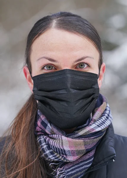 Siyah tek kullanımlık ceketli genç bir kadın. Virüs maskesi, bulanık ağaçlarla kaplı arka plan.