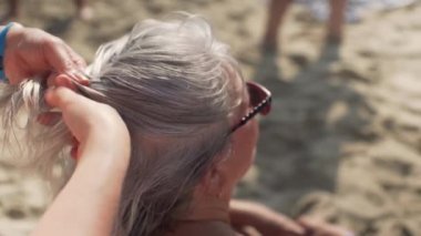 Genç bir kadın yaşlı ve yaşlı annesinin kumsalda dinlenen gri saçlarını örüyor. Kafasına ve ellerine yakın plan, bulanık kum arka plan.