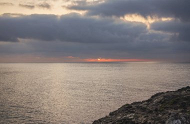 Kızıl güneş denizin üstünde yükseliyor bulutların arkasında, kayalık kıyılarda