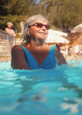 Gri saçlı yaşlı bir kadın, mavi mayo giyiyor, otel havuzunda su aerobiği yapıyor.