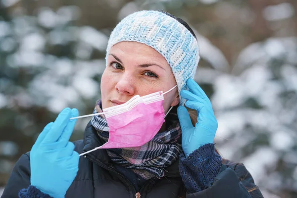 暖かい冬の服の若い女性は 保護手袋を身に着けている 置くか 使い捨ての単回使用ピンクの顔のウイルスの口のマスクを削除 クローズアップ ぼやけた雪に覆われた木の背景 — ストック写真
