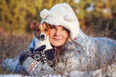 Kışlık ceketli genç bir kadın, karla kaplı zemine uzanmış, elinde teriyer köpeği, bulanık ağaçlar...