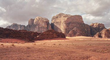 Kızıl kum çölünde Rocky Massifs, birkaç kuru ot kümesi zemin, arka planda bulutlu gökyüzü, tipik Wadi Rum manzarası, Ürdün