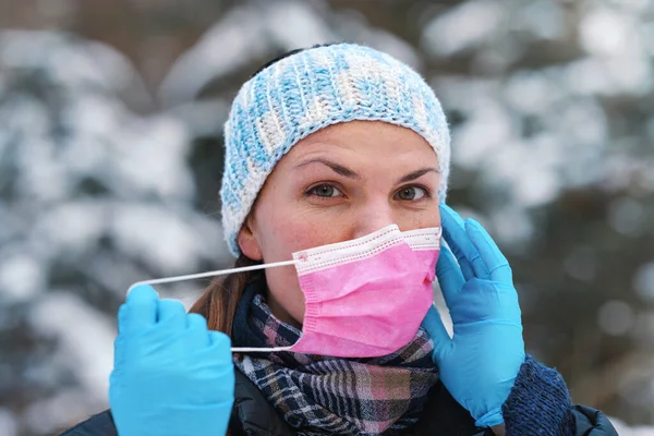暖かい冬の服の若い女性は 保護手袋を身に着けている 置くか 使い捨ての単回使用ピンクの顔のウイルスの口のマスクを削除 クローズアップ ぼやけた雪に覆われた木の背景 — ストック写真