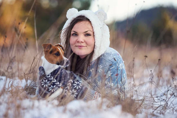 Kışlık ceketli genç bir kadın, karla kaplı zemine uzanmış, elinde teriyer köpeği, bulanık ağaçlar...