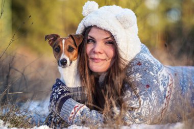 Kışlık ceketli genç bir kadın, karla kaplı zemine uzanmış, elinde teriyer köpeği, bulanık ağaçlar...