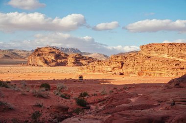 Kırmızı kum çölünde Rocky Massifs, arka planda parlak bulutlu gökyüzü, uzakta küçük araç ve develer - tipik Wadi Rum manzarası, Ürdün