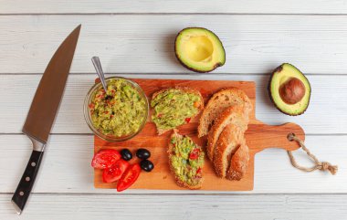 Düz bir fotoğraf, küçük cam kasede yeni hazırlanmış guacamole, ekmek, domates, zeytin ve yanındaki beyaz ahşap masada iki avokado.
