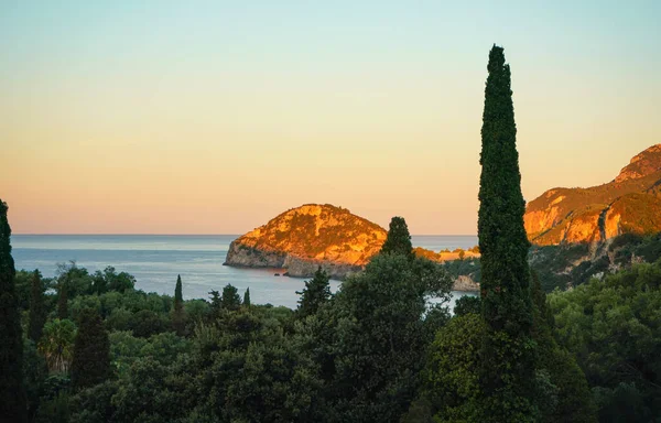 Morning sun shines over hills near Liapades bay in Corfu, tall trees in foreground. Sunrise at sea coast.