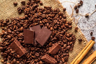 Koyu kavrulmuş aromatik kahve çekirdekleri, koyu ve gözenekli çikolata, gri tarafsız arkaplanda kahverengi bir beze üzerinde tarçın çubukları.