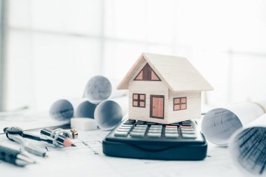 Hesap makinesi, kalem, pusula ve tasarım planı üzerinde mini model ev. Maliyet Mimarı ve Mühendislik kavramı.
