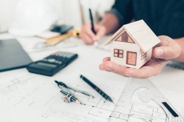 Mimar mühendisin elleri mini ev modelini tutuyor ve taslak üzerinde kalem çizimi yapıyor. Ev planlama ve inşaat konsepti.