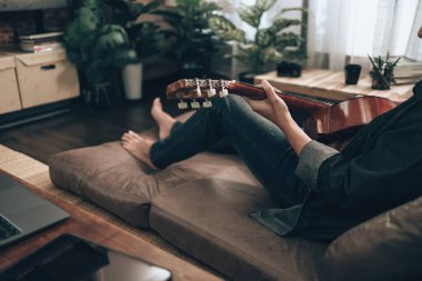 young man relax and playing guitar while sitting on sofa bed in living room at home. Music hobby and practice concept