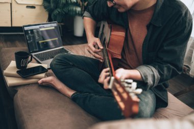 young man relax and playing guitar while sitting on sofa bed in living room at home. Music create melody song, lyrics on laptop and practice concept.