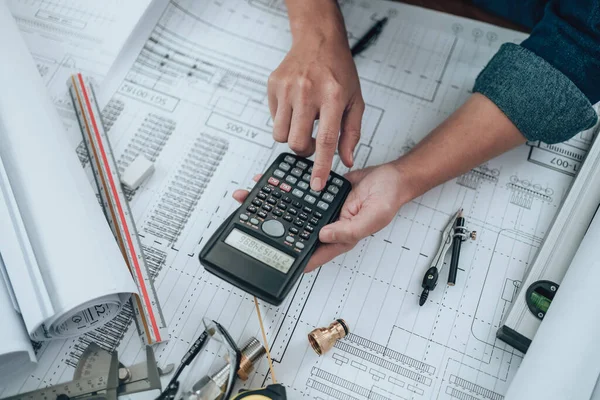 Engineering working with drawings inspection and press calculator on the office desk and triangle ruler, safety glasses, compass, vernier caliper on Blueprint. Engineer, Architect, Industry and factory concept.