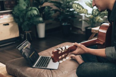 young man relax and playing guitar while sitting on sofa bed in living room at home. Music create melody song, lyrics on laptop and practice concept