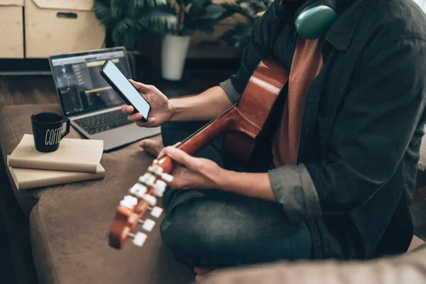 年轻人坐在客厅的沙发床上 在家里用电话搜索音乐 一边放松一边弹吉他 音乐创造旋律歌曲 笔记本电脑上的歌词和实践概念 — 图库照片