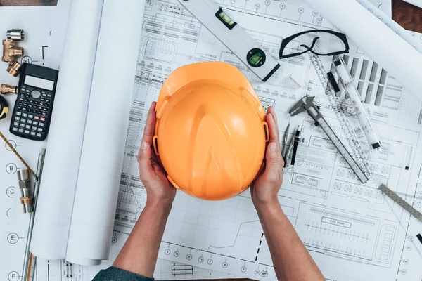 Engineering holding safety helmet  with drawings inspection on the office desk and Calculator, triangle ruler, safety glasses, compass, vernier caliper on Blueprint. Engineer, Architect, Industry and factory concept.