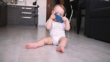 Sevimli bebek telefonu elinde tutuyor. Çocuk cep telefonuyla oynuyor, onu inceliyor, ellerini çekiştiriyor ve akıllı telefon yemeye çalışıyor, ağzına koyuyor.