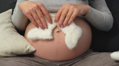 Mutlu hamile kadın evdeki kanepede otururken bebek kıyafetlerine şefkatle bakıyor. Müstakbel annenin doğumdan önce doğmamış bebek için alışveriş yapması. Manzarayı kapat. Yüksek